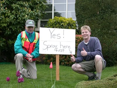 two men by on a residential lawn near a sign that says "Yes! Spray here first"